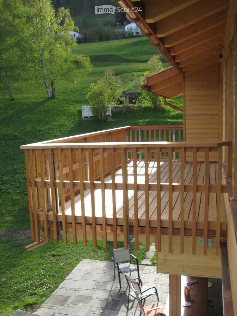 Wooden balcony with railing, overlooking a green, hilly landscape. Two chairs placed on a paved area below the balcony.