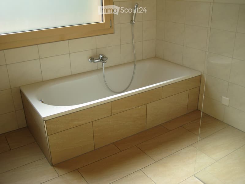 Modern bathroom with a corner bathtub, tiled walls, wooden floor, and a window above the tub.