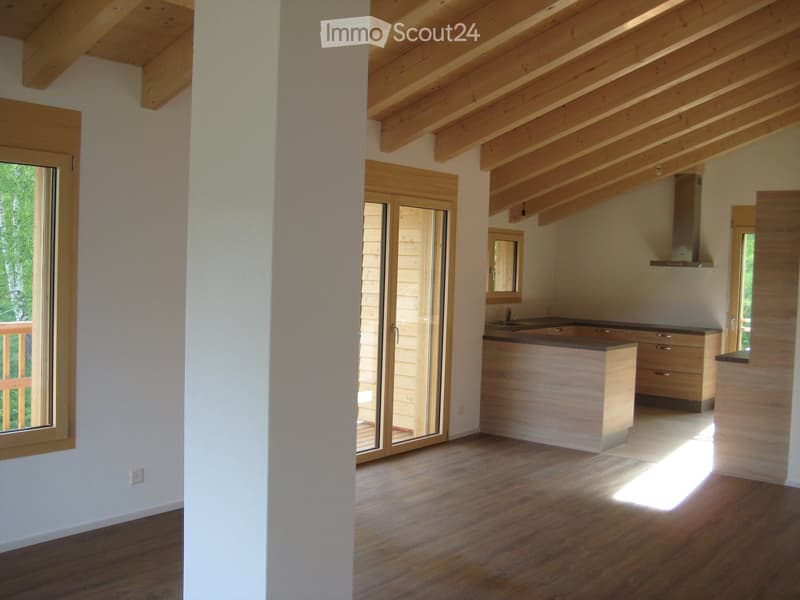 Open-plan space with wooden flooring, exposed wooden beams on the ceiling, large windows, and a kitchen area with cabinets and a sink.