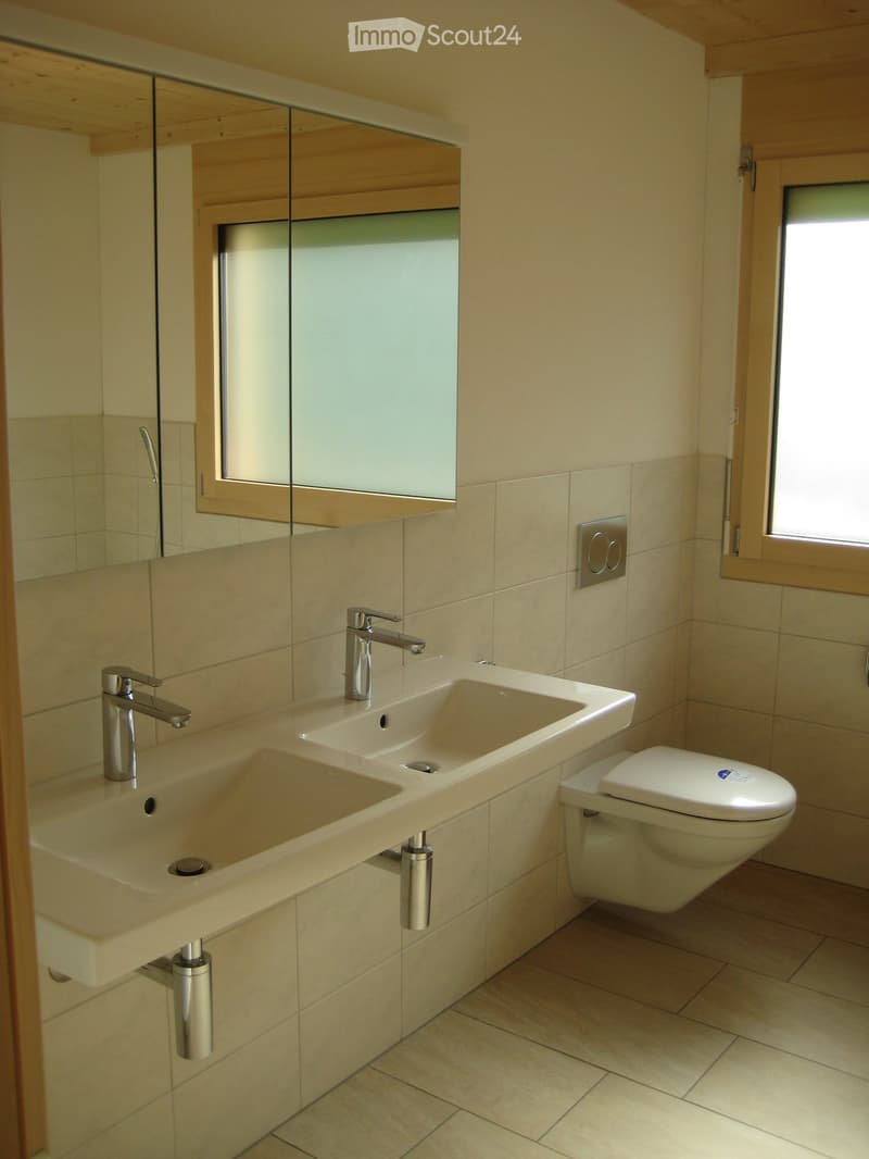 Modern bathroom with a double sink, chrome faucets, and a toilet. Light-colored tiles and wooden accents.