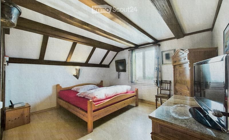 Cozy attic bedroom with wooden beams, a single bed with red bedding, a small wooden chair, a wardrobe, and a table with a screen.