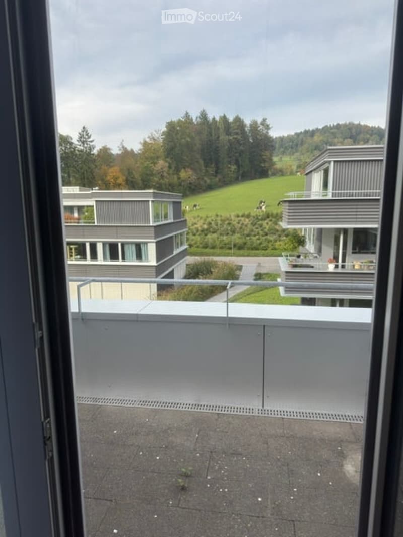 Balcony with a modern glass railing, overlooking a green landscape with trees and a modern building in the background.