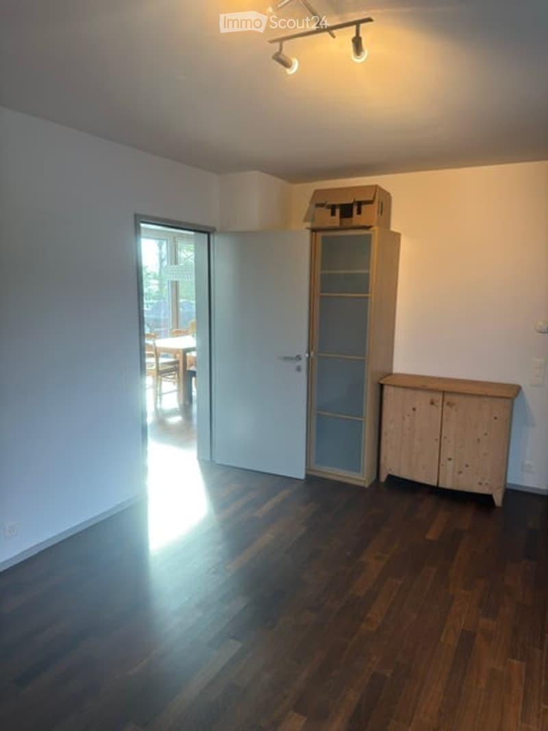 Empty room with wooden flooring, white walls, and a glass-fronted cabinet. A door leads to another room with a dining table and chairs visible in the background.