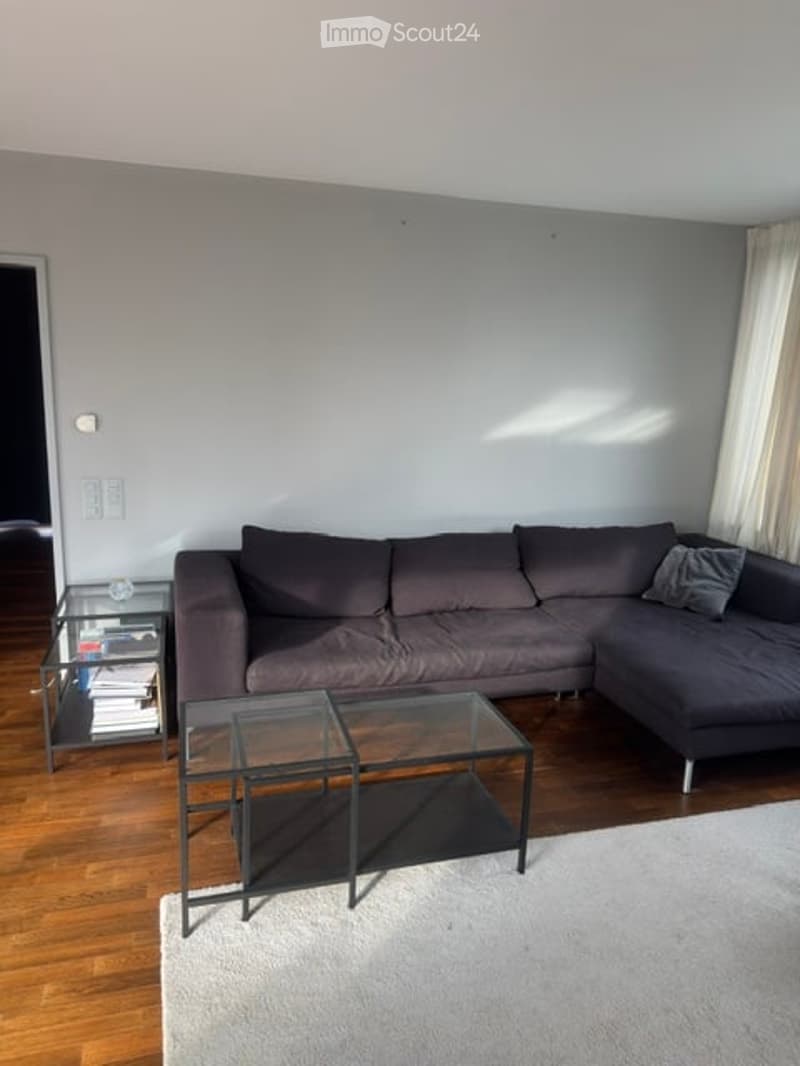 Modern living room with grey sectional sofa, glass coffee table, side table, and books on a shelf. Light grey walls and wooden flooring.
