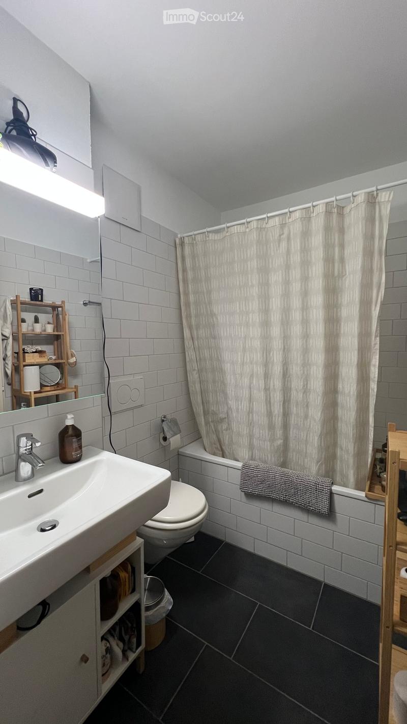 Modern bathroom with white tiles, a sink, toilet, bathtub, and a shower curtain. Includes a wooden shelf with various items and a towel on the edge of the bathtub.