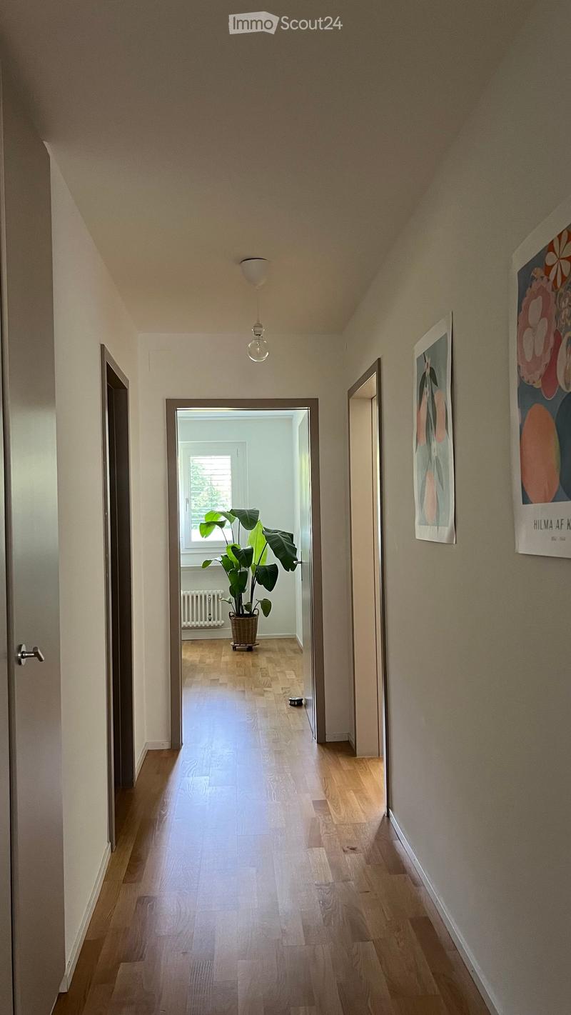 A narrow hallway with wooden flooring, light-colored walls, and a hanging light fixture. Two framed artworks are displayed on the right wall, and an open doorway leads to another room with a potted plant visible.