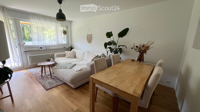 Bright living room with a white sectional sofa, wooden dining table, and hanging plants. Features large windows with sheer curtains and a pendant light.