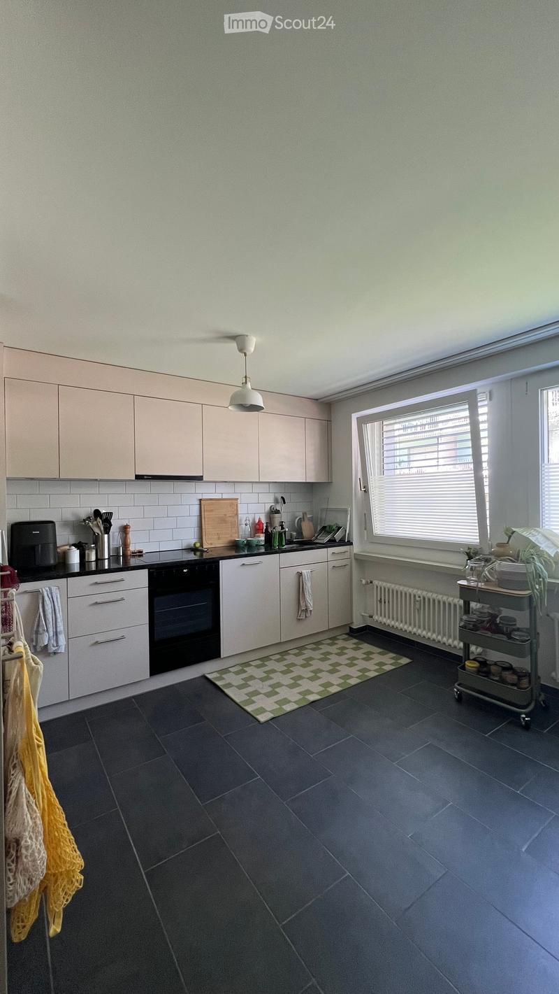 Modern kitchen with light-colored cabinets, dark countertops, and a dark tiled floor. Features include an oven, sink, coffee maker, and a small cart with jars.