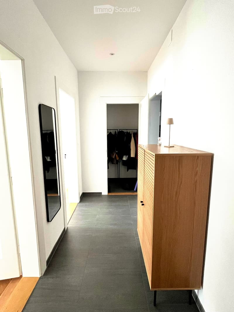 A modern, minimalist hallway with white walls, grey tiled floor, and a wooden sideboard with a lamp on top. A closet with hanging clothes is visible in the background.