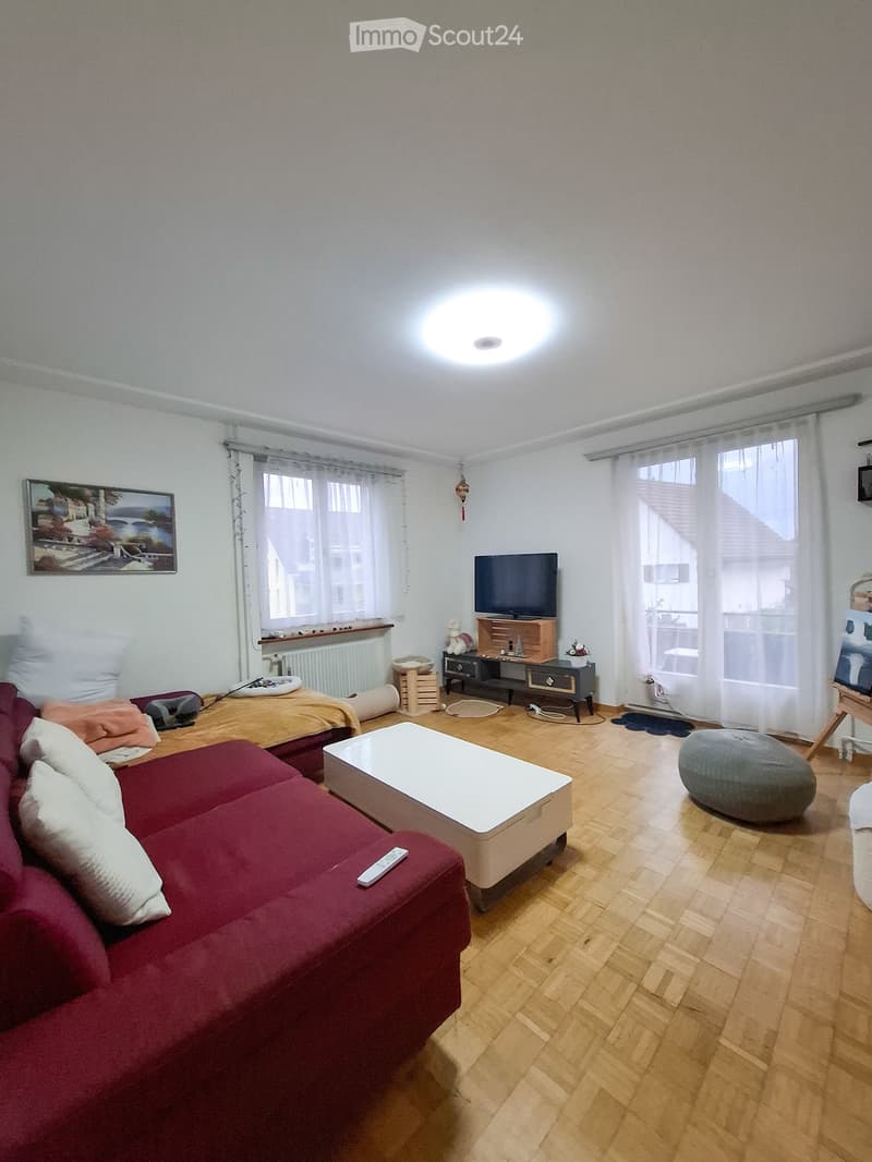 A modern living room with a red sofa, white coffee table, grey ottoman, television, and artwork on the wall. The room features wooden flooring and large windows with white curtains.