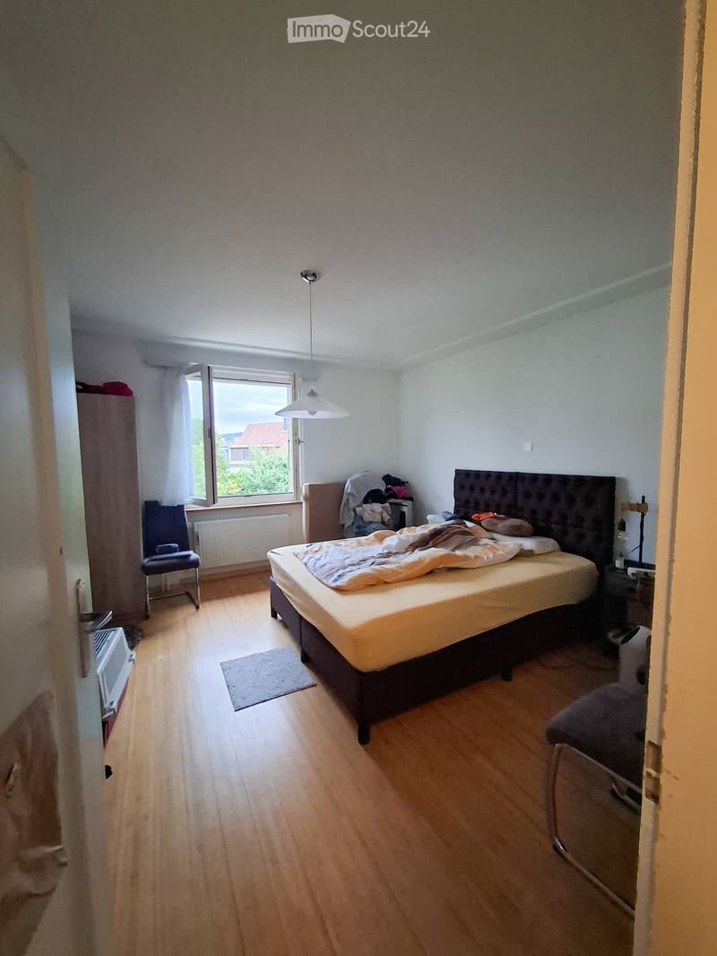 Bedroom with wooden flooring, a bed with a dark headboard, a chair, a wardrobe, and a radiator near the window.
