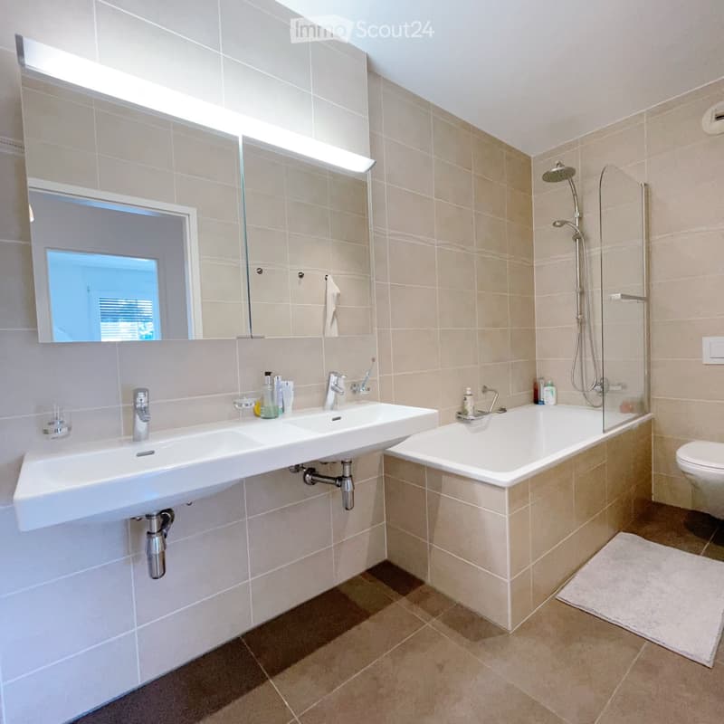 Modern bathroom with beige tiles, double sink, bathtub, toilet, and showerhead. Includes a large mirror, towel, and toiletries.