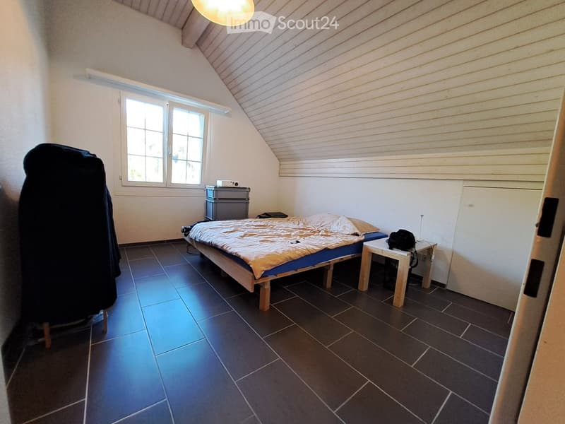 Bedroom with a bed, nightstand, small table, chair, and wardrobe. The room has a slanted ceiling and large windows.