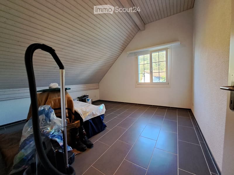Empty room with tiled floor, slanted wooden ceiling, and a window with white frame. A vacuum cleaner is partially visible in the foreground, and some items are stacked near the window.