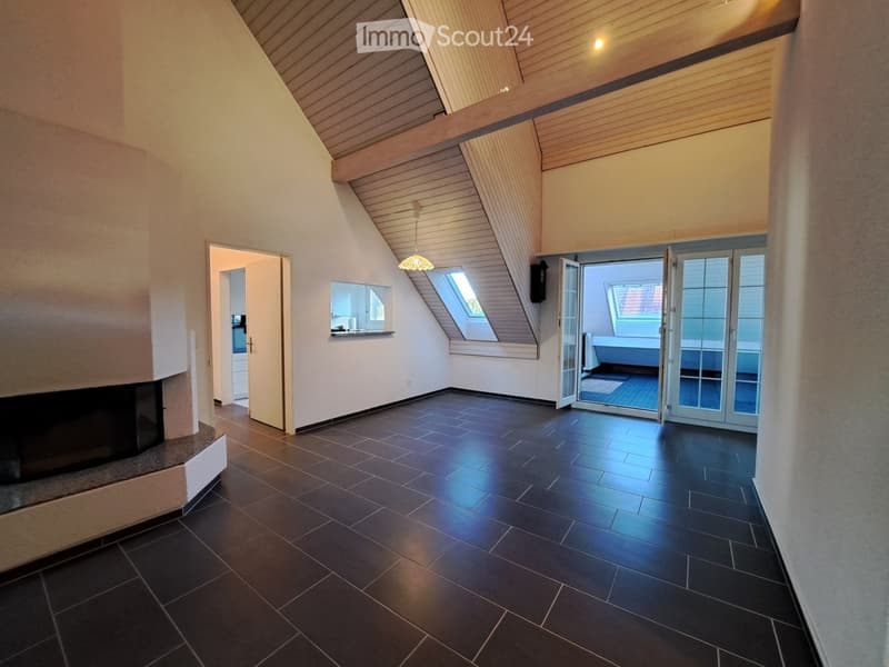 An empty room with dark tiled flooring, white walls, and a slanted wooden ceiling. Features include a fireplace on the left, a doorway leading to another room, and large glass doors opening to a balcony or outdoor area.