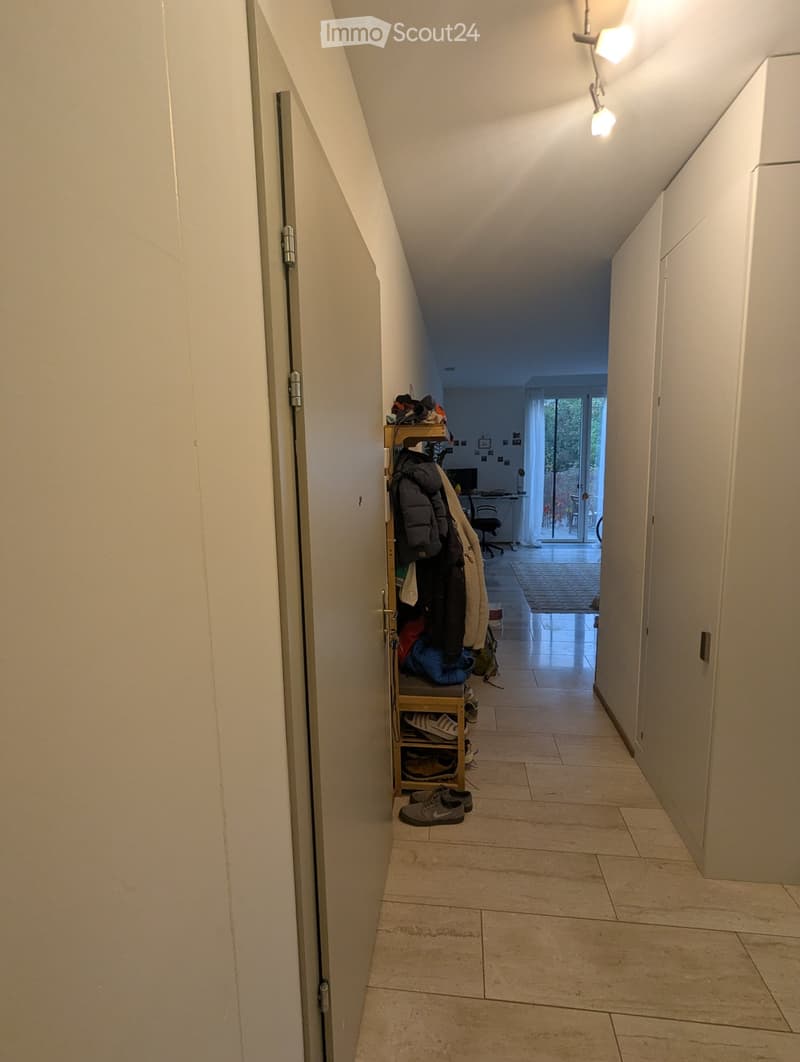 A narrow hallway with tiled flooring, white walls, and built-in storage shelves holding shoes and jackets. The hallway leads to a room with a desk and a sliding glass door to an outdoor area.