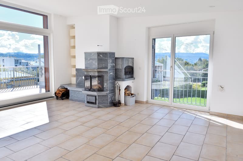 Bright living room with large windows and sliding glass doors leading to a balcony, featuring a stone fireplace with wood storage and tools.