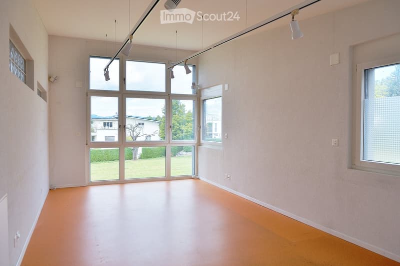 Empty room with light wooden flooring, white walls, large windows, and track lighting. No furniture or equipment present.
