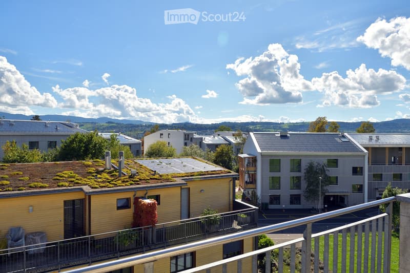 View of a residential complex with modern buildings, green roofs, and surrounding greenery under a clear blue sky.