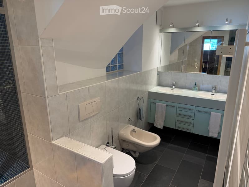 Modern bathroom with a toilet, bidet, double sink, large mirror, and light blue cabinets. The walls are tiled in light gray, and the floor is dark gray.