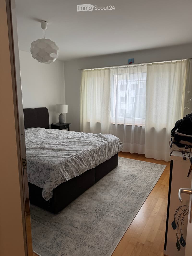 A modern bedroom with a large bed, nightstand, lamp, and a large window with curtains. The room features wooden flooring and a decorative rug.