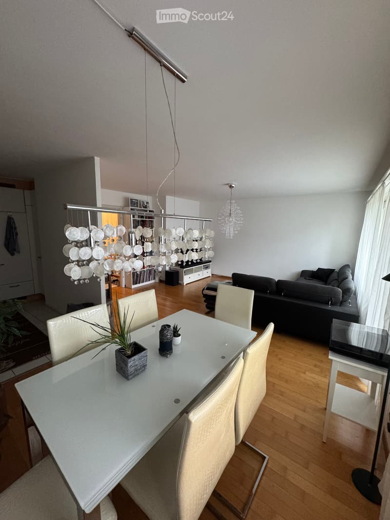 Modern dining area with a white glass dining table, four white chairs, and a small plant on the table. A gray sofa and a black coffee table are visible in the background, along with a chandelier and a bookshelf.