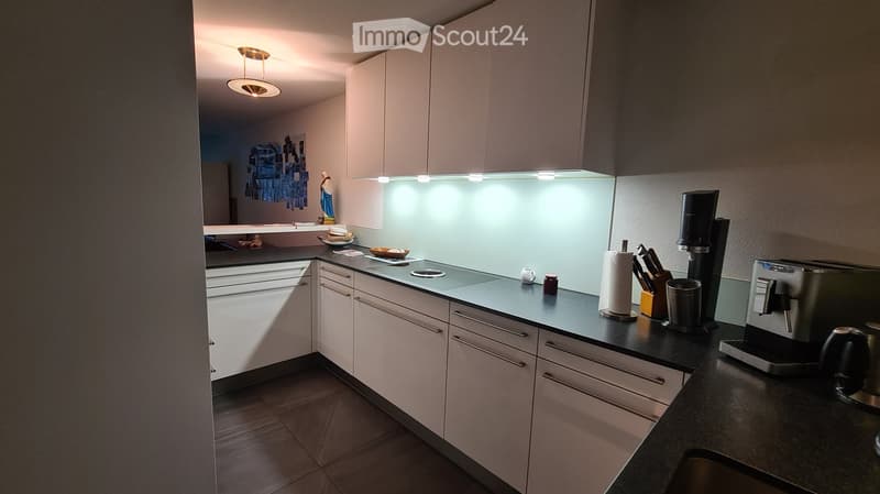 Modern kitchen with white cabinets, black countertops, and various appliances including a coffee maker and toaster. The space features a sleek design with integrated lighting and a clean, organized layout.