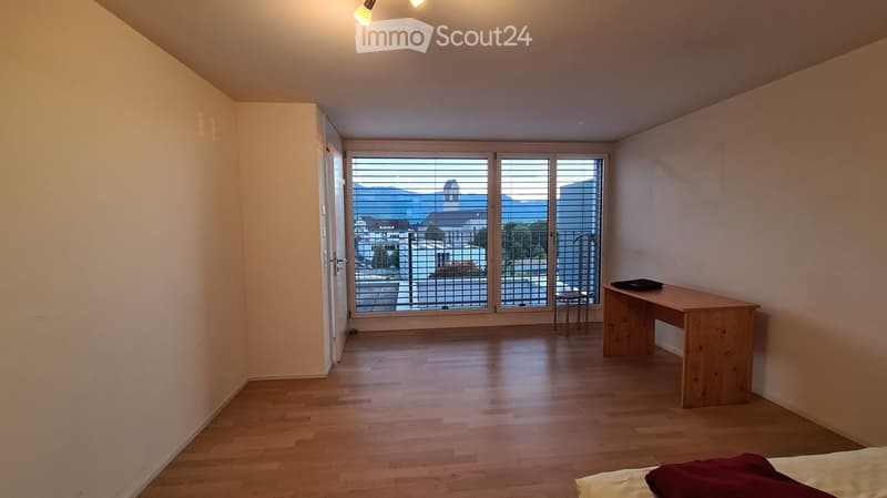 Bright, unfurnished room with wooden flooring, large windows offering a scenic view, a desk, a stool, and a bed with pillows.