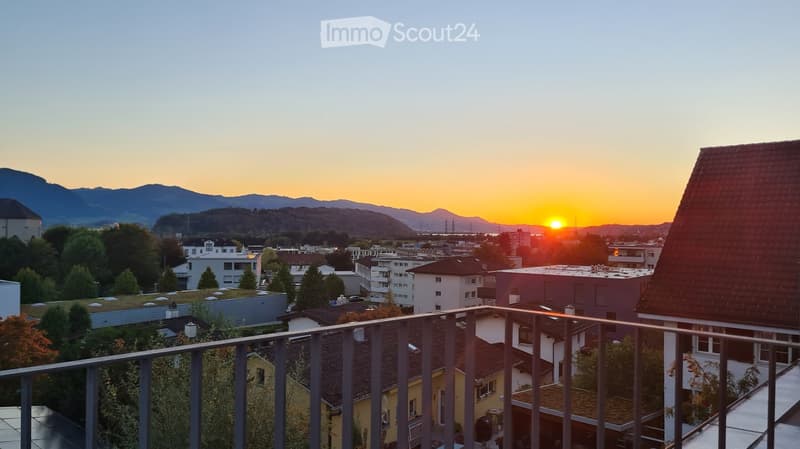 Balcony overlooking a cityscape at sunset, with visible rooftops, trees, and distant mountains.