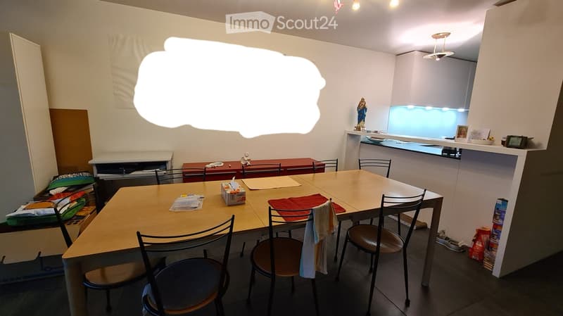 A furnished room with a dining table and chairs, a kitchen area with a counter, and various items on the counter and shelves.