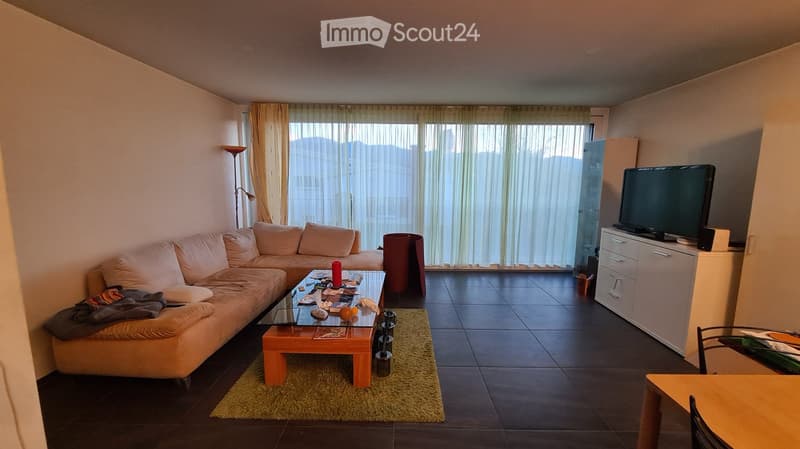 Living room with a beige sofa, coffee table, chair, and television. Large windows with curtains provide a view of the city.