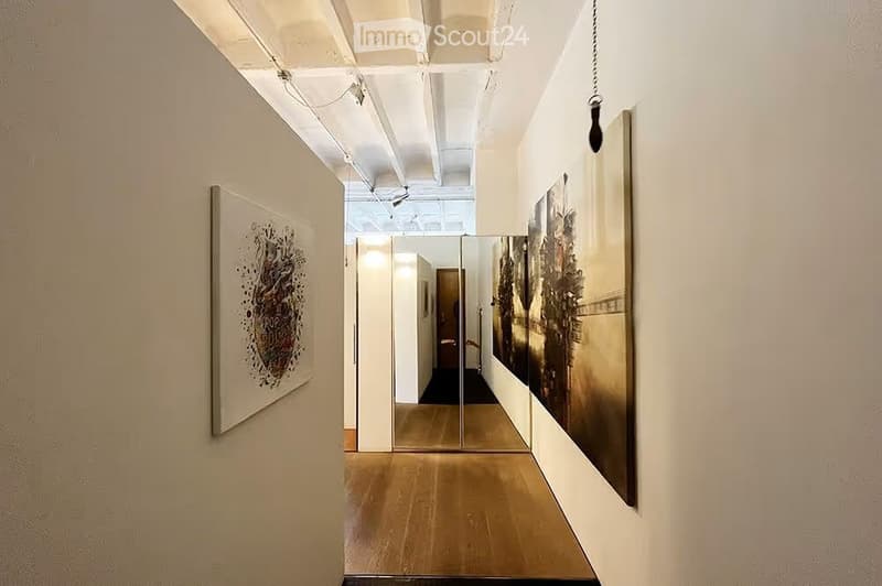 Modern hallway with wooden flooring, white walls, and large framed artwork on both sides. Exposed ceiling with industrial-style lighting fixtures.