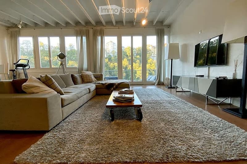 Modern living room with large windows, a sectional sofa, coffee table, floor lamp, and a television mounted on the wall. An exercise bike and a globe on a tripod are also visible.