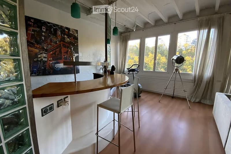 Modern, open-plan room with a bar counter, two stools, large windows with curtains, and a tripod with a camera in the corner.
