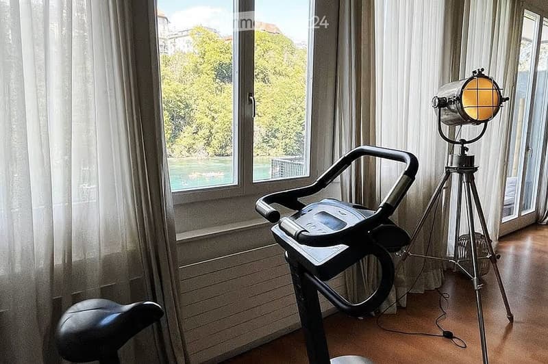 A room with a treadmill and a vintage-style lamp on a tripod stand, large windows with sheer curtains, and a view of greenery outside.
