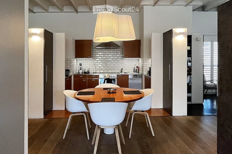 Modern kitchen and dining area with a round wooden dining table, white chairs, and various kitchen appliances including an oven, dishwasher, and refrigerator. The space features wooden flooring and white walls.