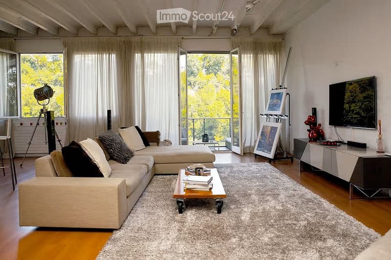 Modern living room with large windows and a view, featuring a beige sectional sofa, a white coffee table with books, a television, and decorative items.