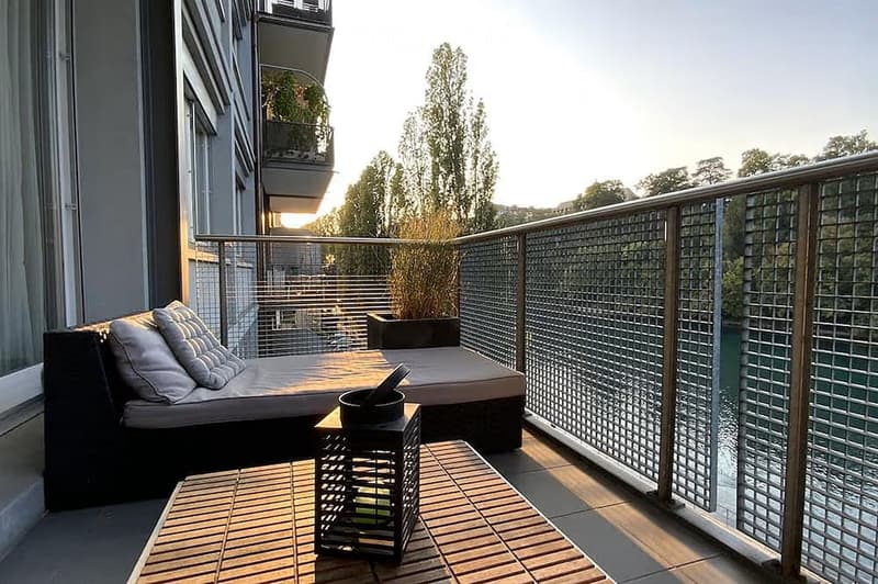 Modern balcony with wooden decking, glass railing, and a comfortable sofa with cushions, a small table with a pot, and a potted plant.