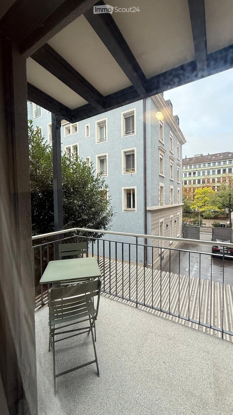 Balcony with a small table and two chairs, overlooking a street with buildings and greenery.