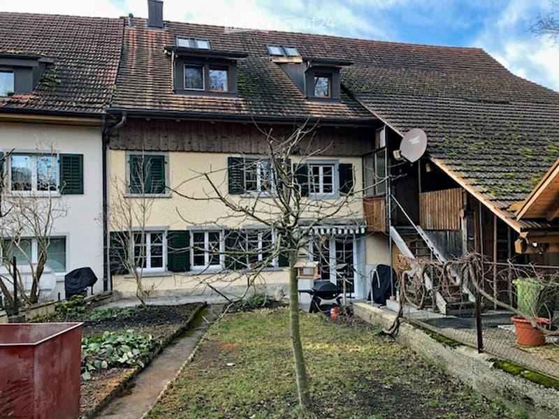 Two story house, brown roof, large windows, satellite dish, stairs leading to the entrance, fence, green potted plant, grass, small tree, small garden, barbecue.