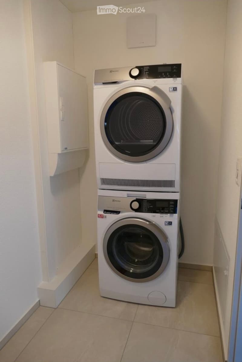 A compact bathroom with a stacked washing machine and dryer unit, beige tiled floor, and white walls.