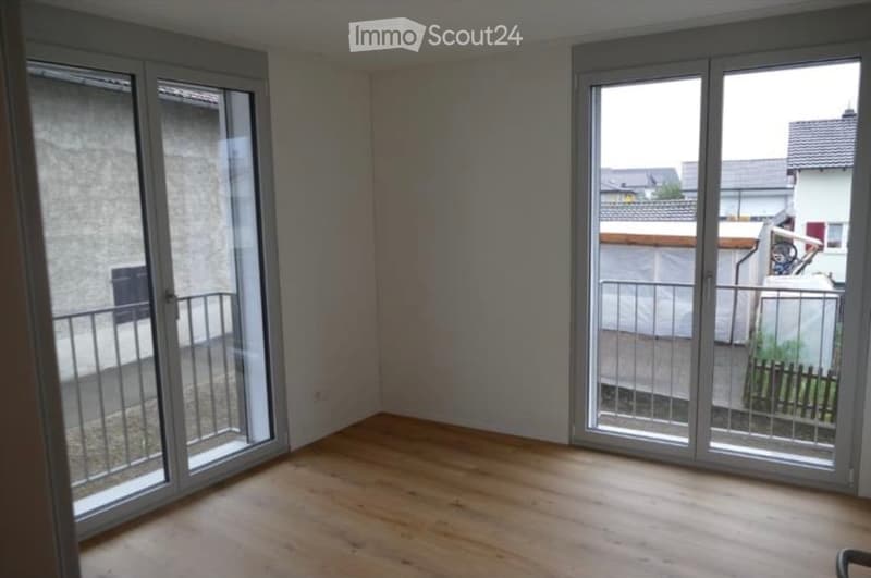 Empty room with wooden flooring, large sliding glass doors leading to a balcony, and a view of neighboring buildings.