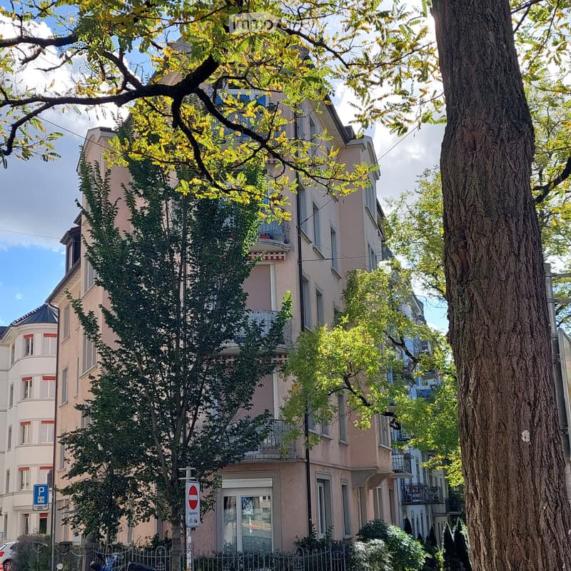 Multi-story residential building with balconies, surrounded by trees and greenery.