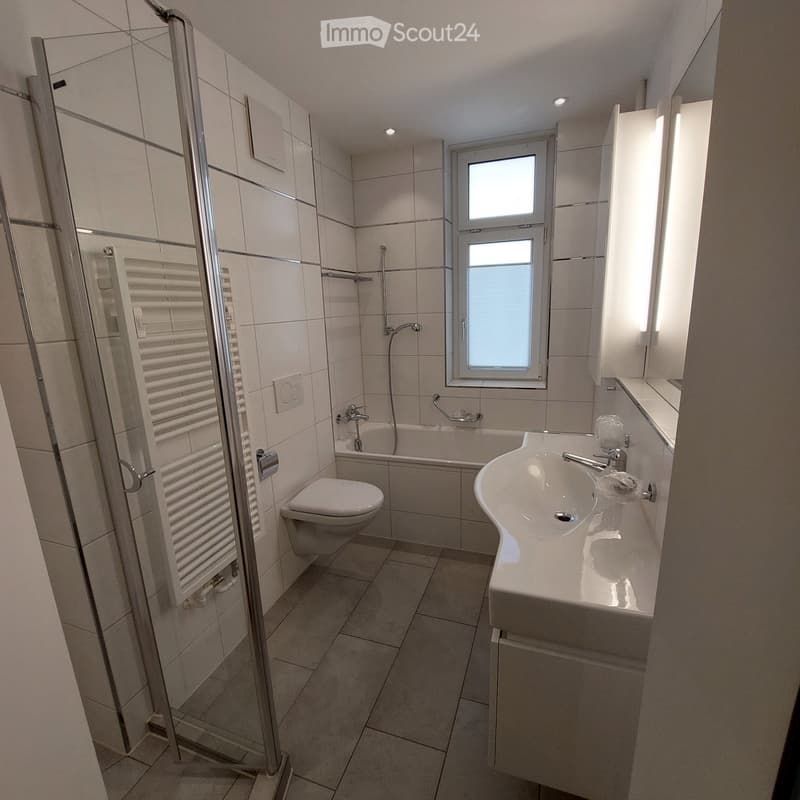 Modern bathroom with white tiles, a bathtub, toilet, sink, and a glass-enclosed shower area.