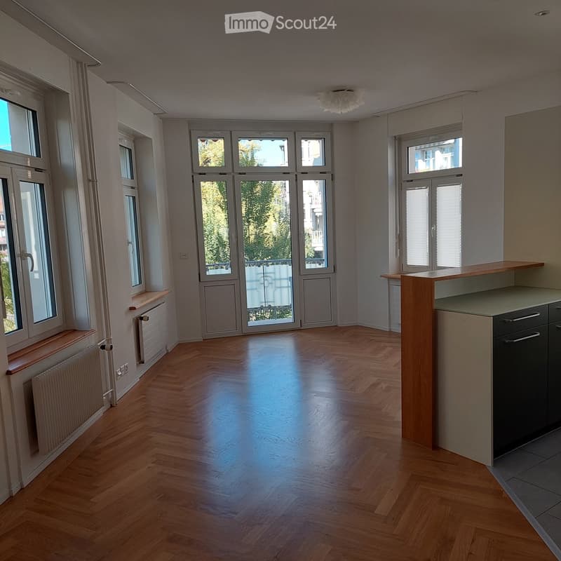 Empty room with wooden flooring, large windows, and a small kitchen area with cabinets and drawers.