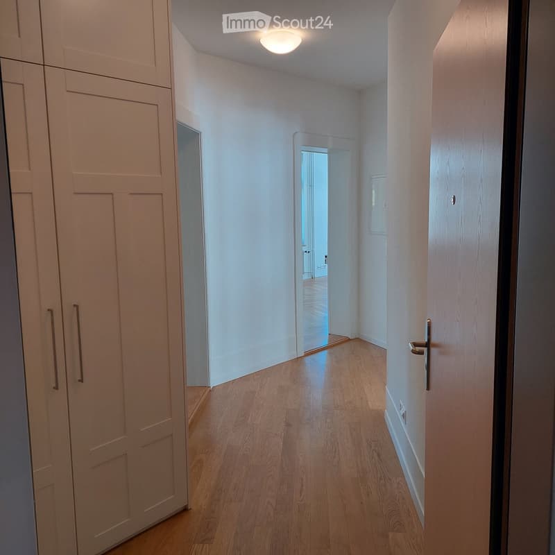 A narrow hallway with light wooden flooring, white walls, and a recessed ceiling light. Doors are visible on both sides, with one door slightly ajar, revealing a glimpse of another room.
