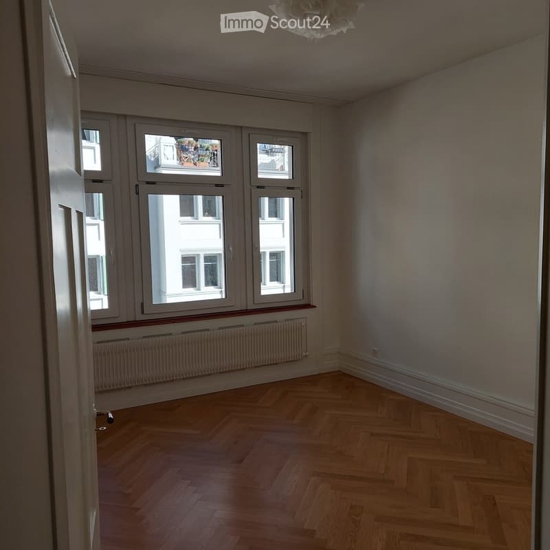 Empty room with wooden herringbone flooring, large windows, radiator, and white walls.