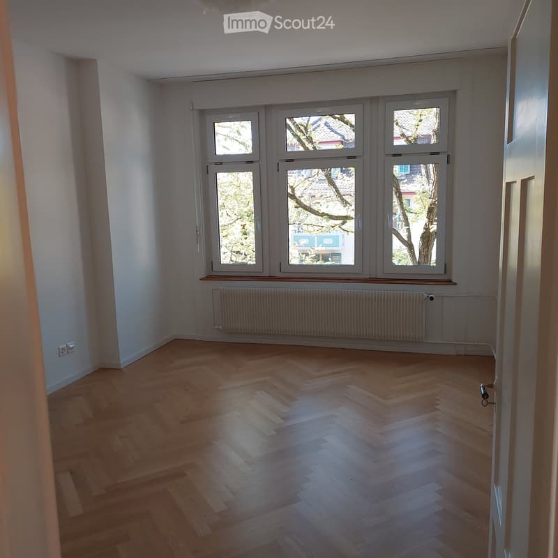 Empty room with wooden flooring, large windows, radiator, and visible electrical outlet.