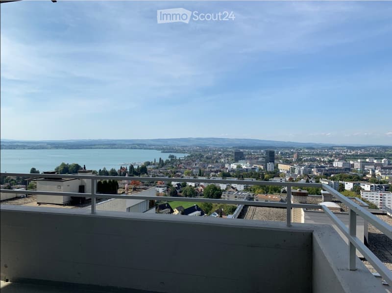Balcony with metal railing overlooking a cityscape, lake, and distant hills under a clear sky.