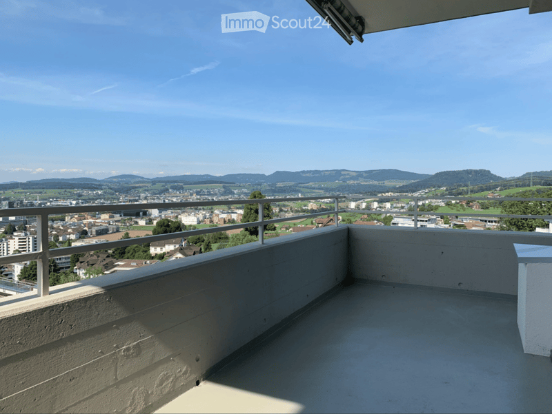 Balcony with metal railing, overlooking a cityscape and distant mountains under a clear blue sky.